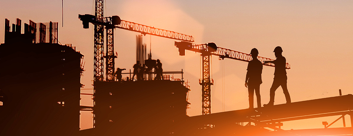 Silhouette of 2 builders pointing to other builders working on a construction site, cranes are in the background. Sun setting in the corner. 